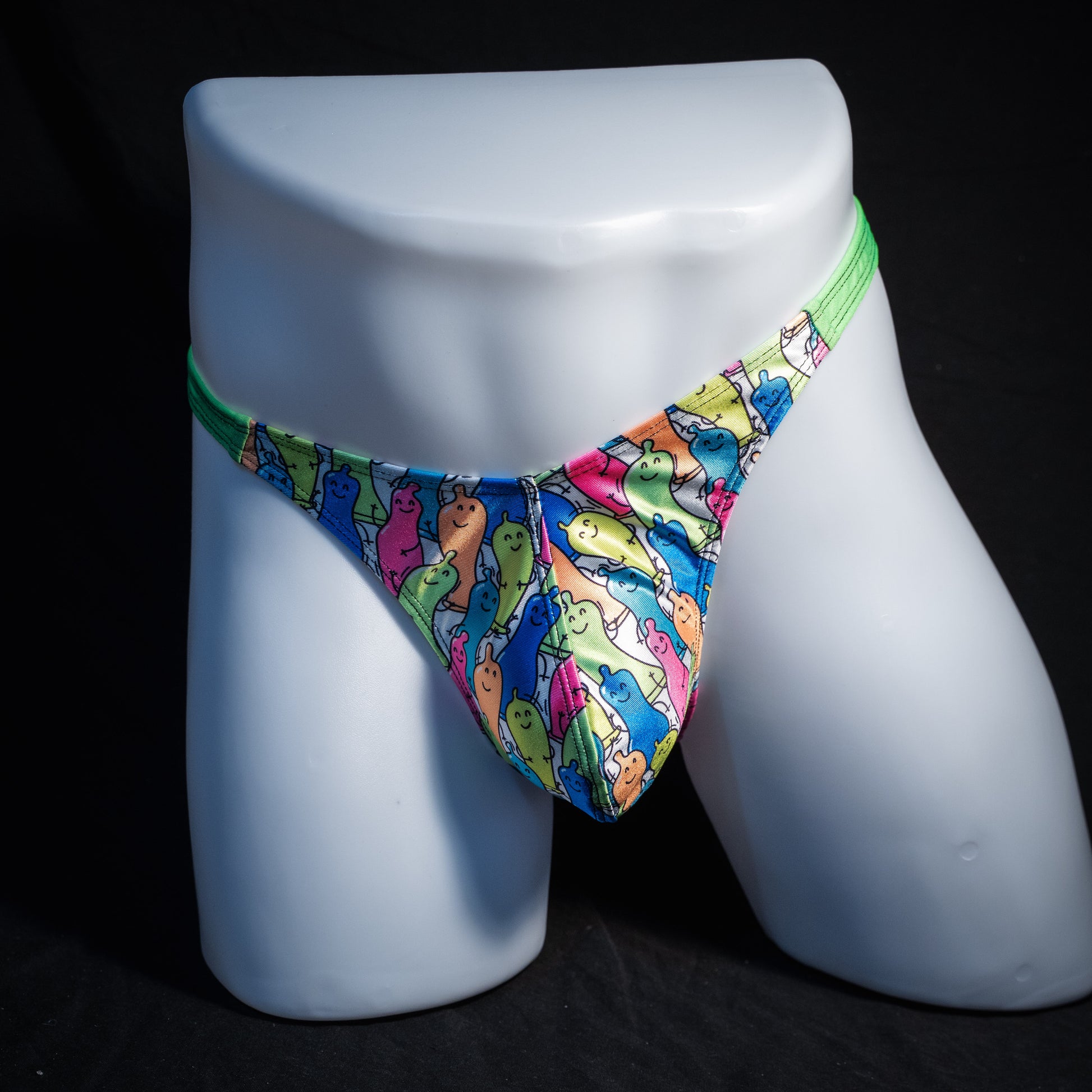 A men's low-rise thong brief with a vibrant multicolor cartoon dinosaur pattern, displayed on a white mannequin leg against a black background.