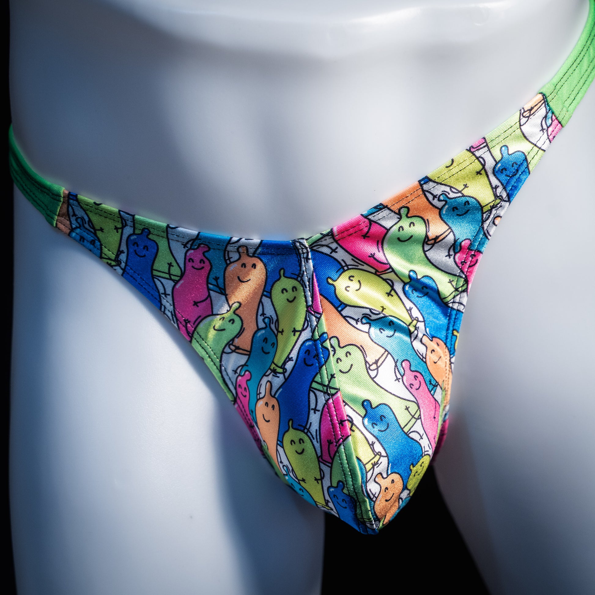 A close-up of a colorful thong-style bikini bottom displayed on a white mannequin torso. The fabric has an allover cartoon ghost print in bright blue, green, pink, yellow, and orange on a white base, with a solid neon green waistband.