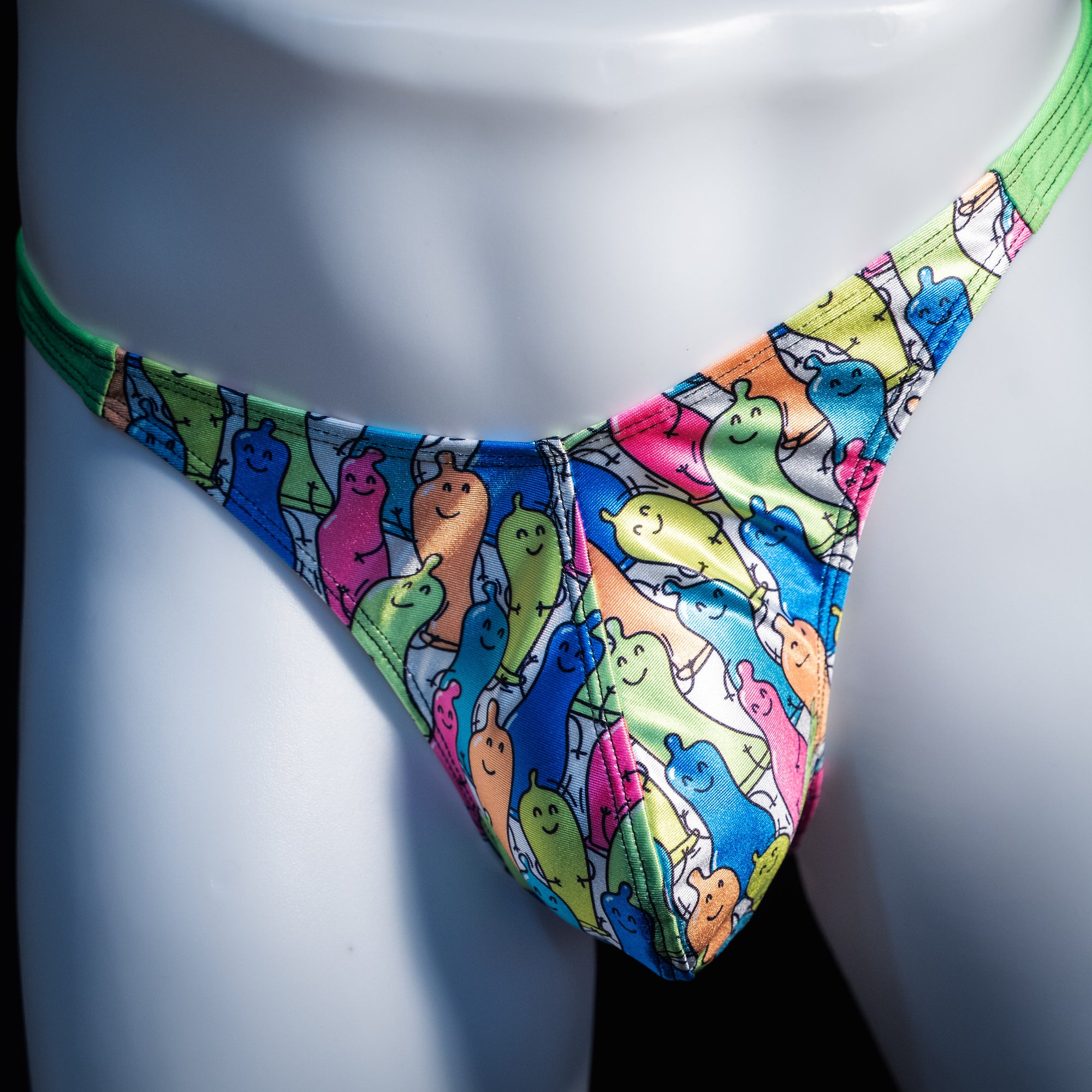 A close-up of a colorful thong-style bikini bottom displayed on a white mannequin torso. The fabric has an allover cartoon ghost print in bright blue, green, pink, yellow, and orange on a white base, with a solid neon green waistband.