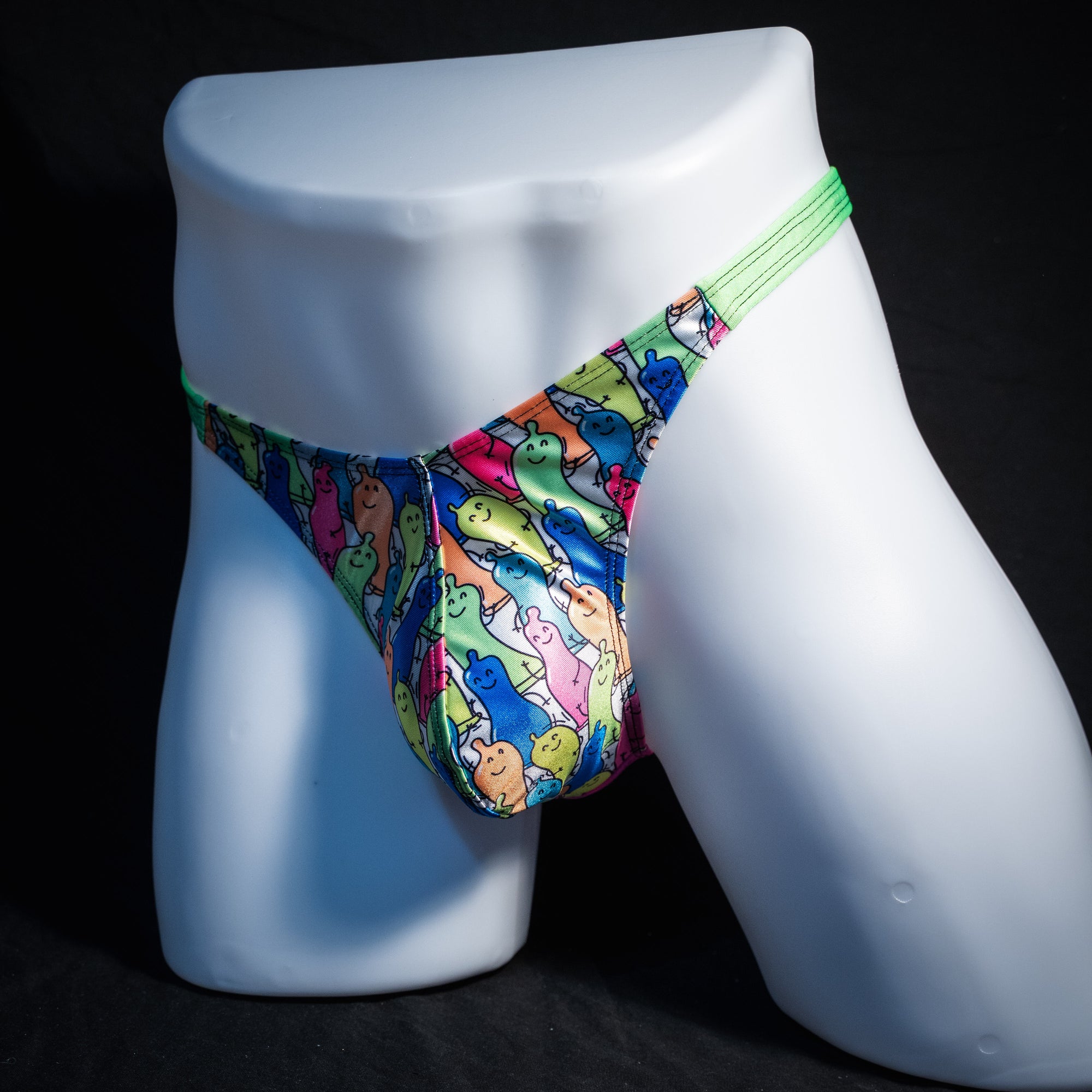 A pair of men's thong underwear with a colorful cartoon dinosaur pattern and a bright green waistband, displayed on a white mannequin against a black background.