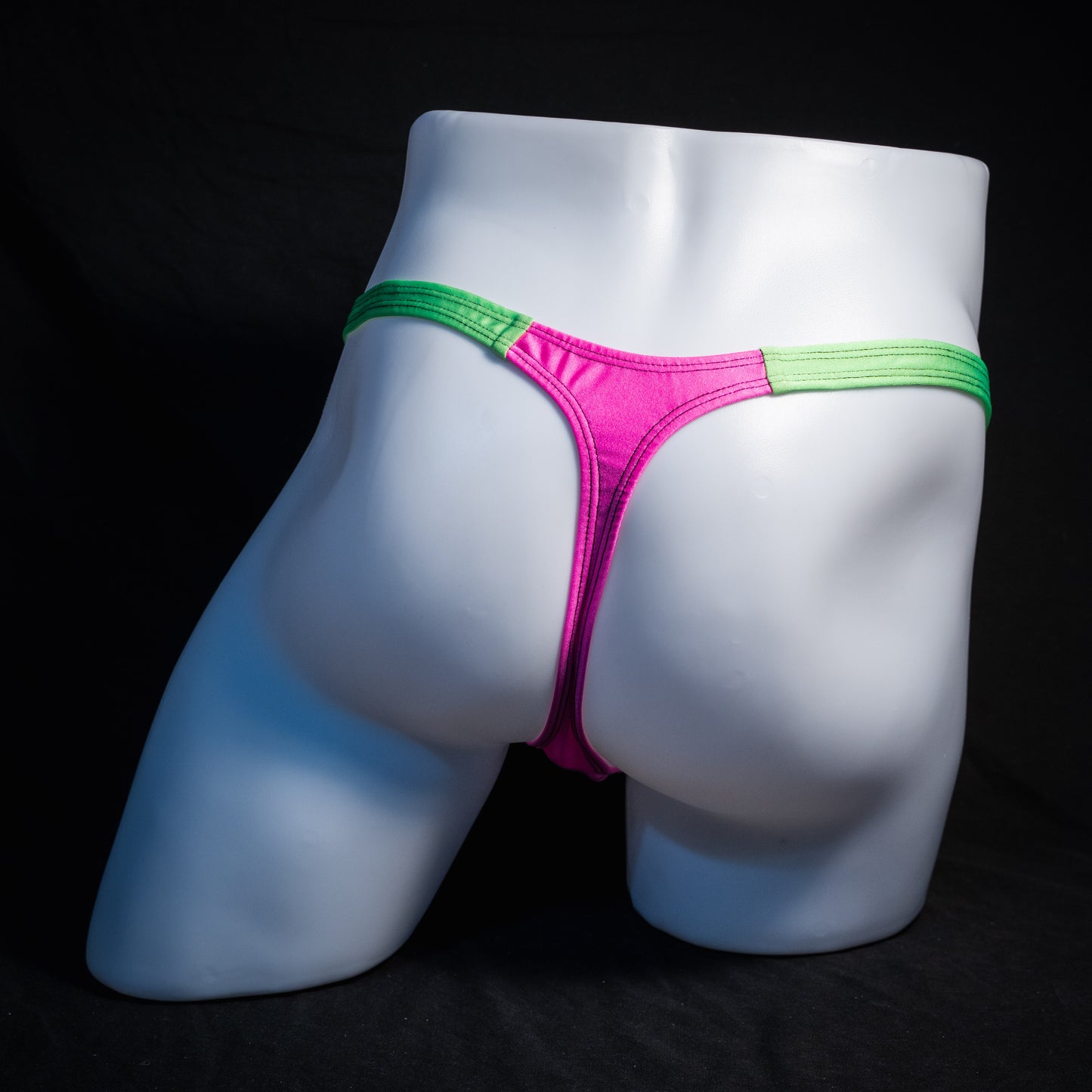 A bright pink jockstrap with lime green elastic waistband and leg straps, displayed on a white mannequin torso against a black background.
