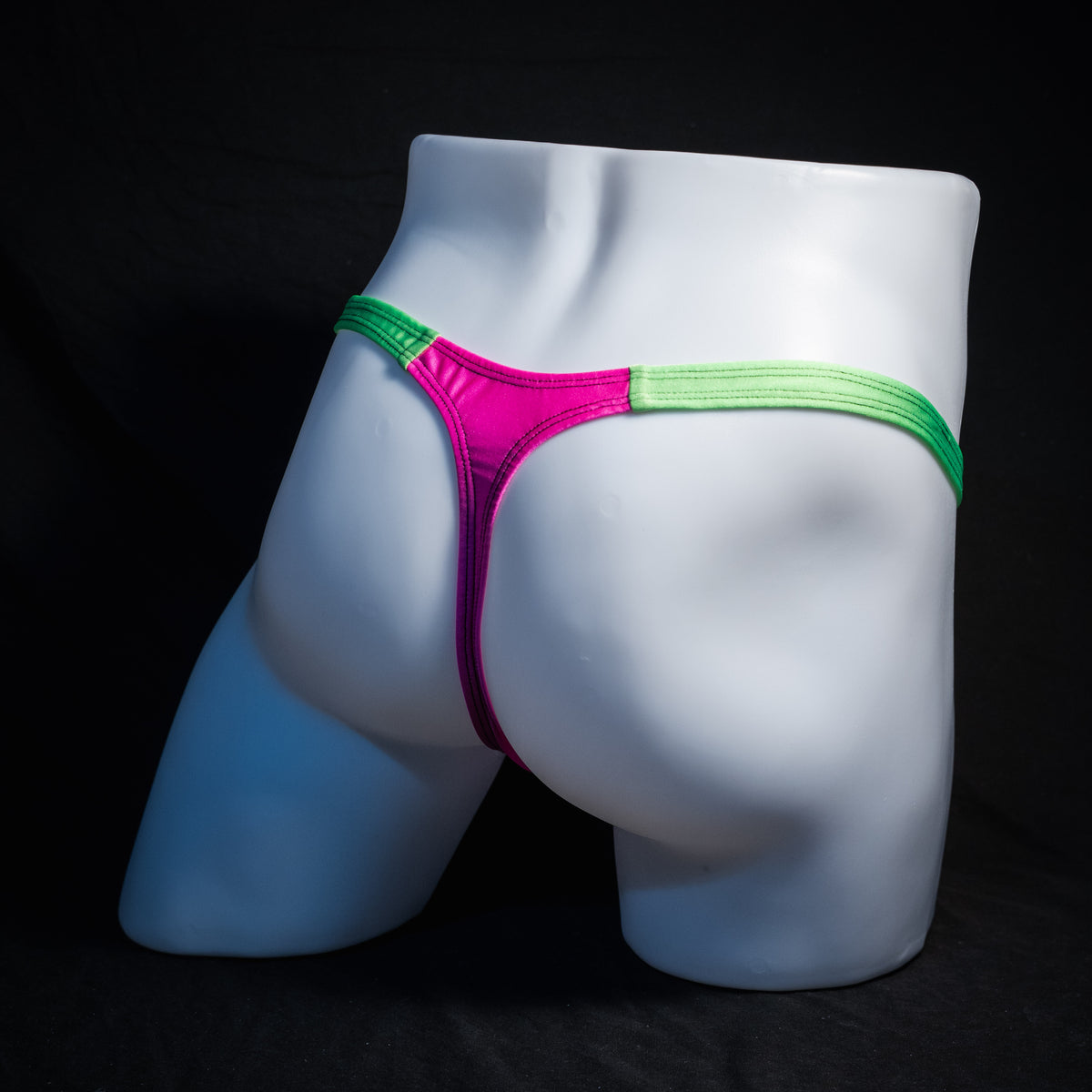 A close-up studio shot of a white mannequin lower torso wearing a thong-style underwear bottom with bright pink fabric and neon green trim and waistband, set against a black background.